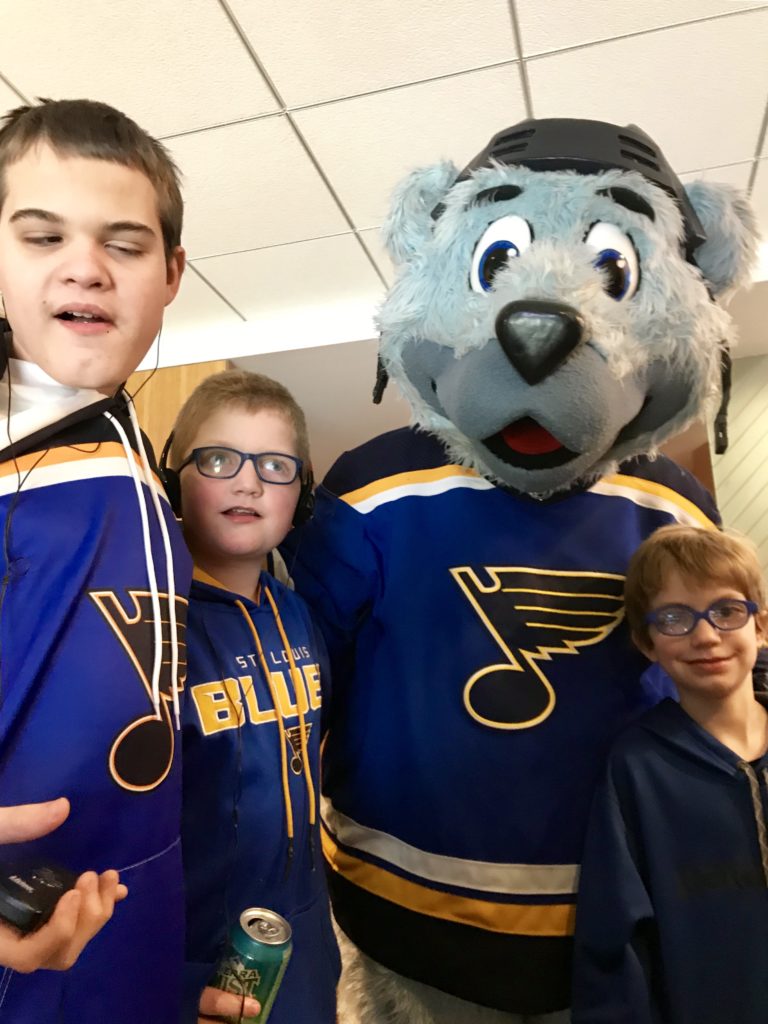 Three children wearing Blues shirts and audio description headsets stand with Louie, the large blue bear mascot of the St. Louis Blues.