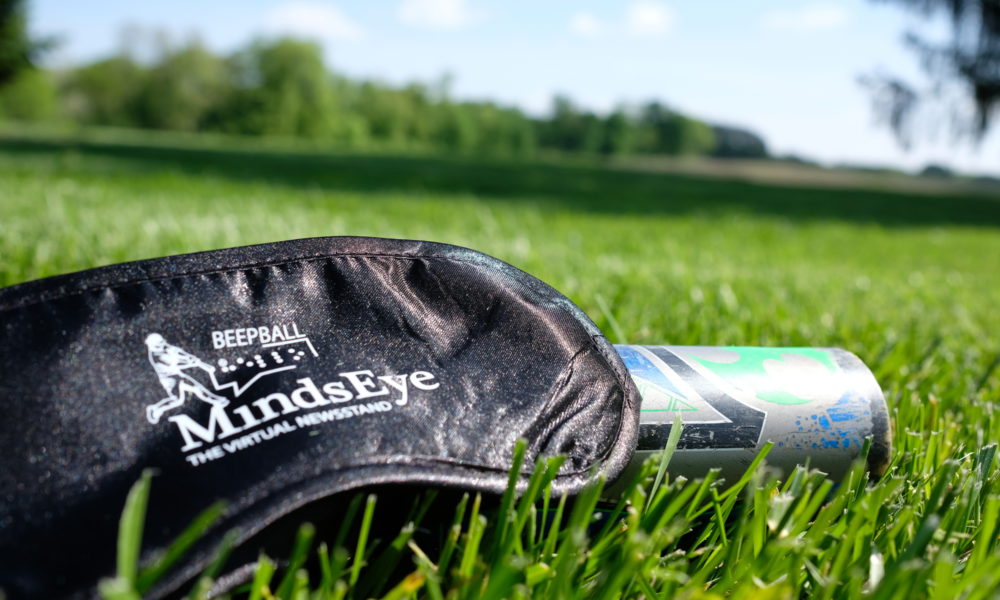 A MindsEye BeepBall mask rests on an aluminum bat in the grass.