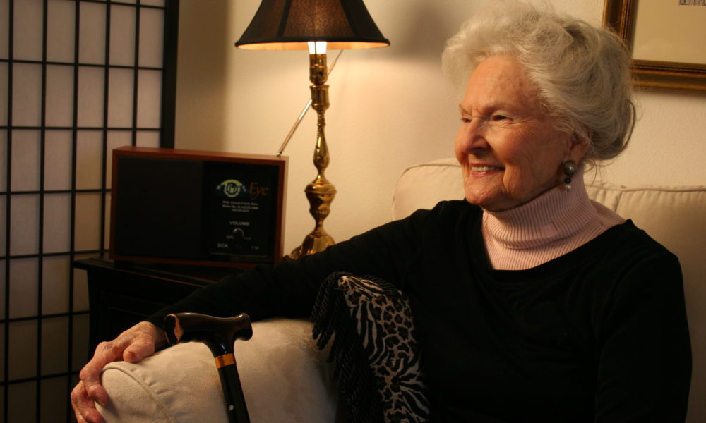 A woman with gray hair sits in front of a MindsEye radio