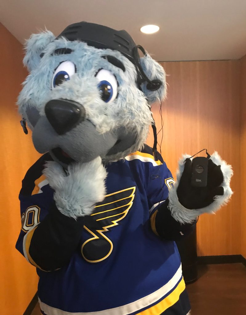 Louie, the Blues Mascot, holds an audio description device with a headset over one large ear.