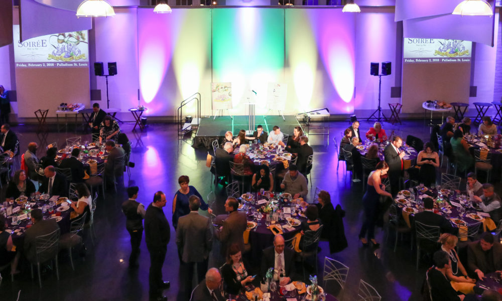 A stage is lit with purple, green, and gold lights at the front of a room filled with elegantly dressed guests seated at round tables.