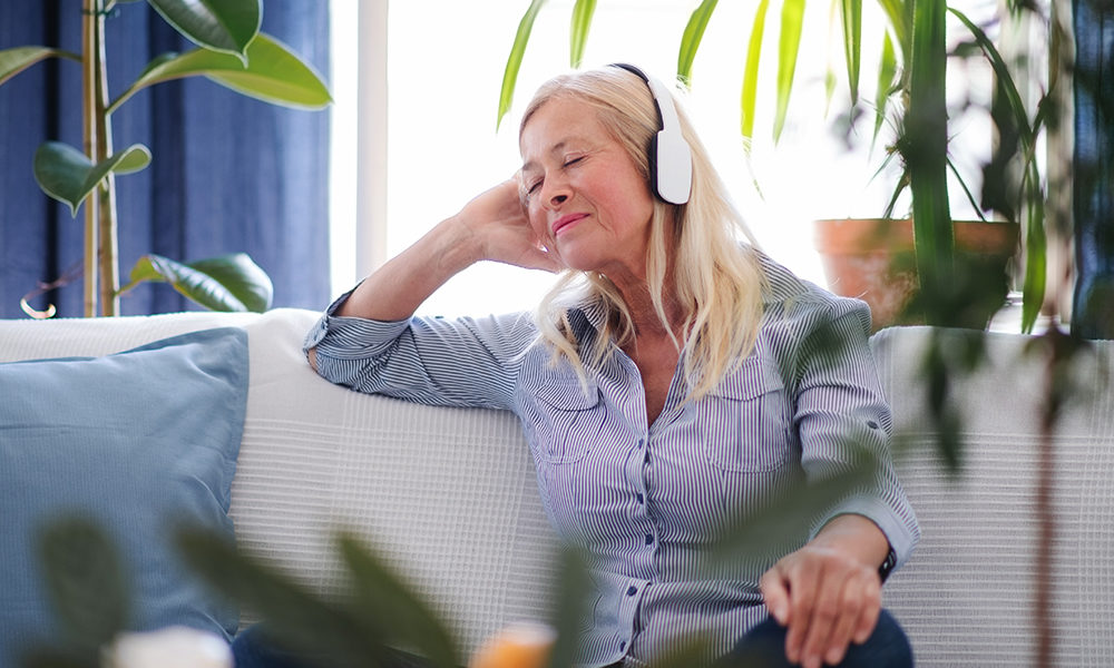 A blond, senior woman wearing headphones while sitting on a couch