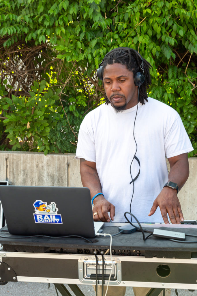 RailDurrty aka DJ ASC stands behind his DJ setup wearing headphones