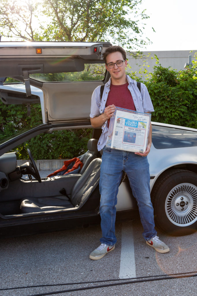A young man dressed like Marty McFly poses in front of his Delorean holding a mockup of an issue of USA Today from the film Back to the Future 2