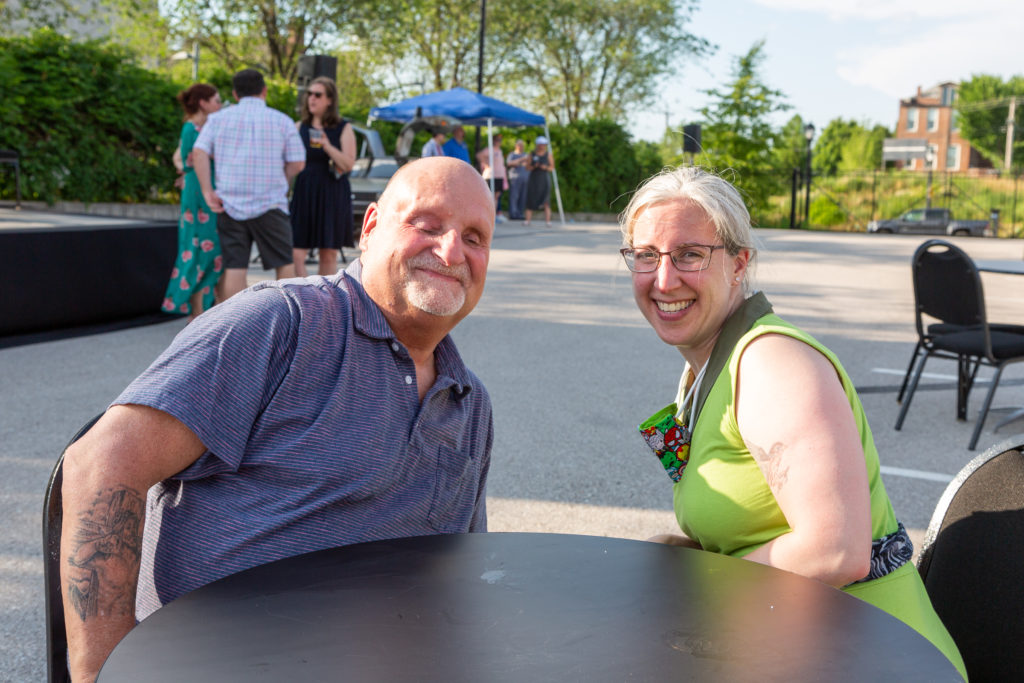 Magan Harms and a friend sit at a table in a parking lot smiling at the camera