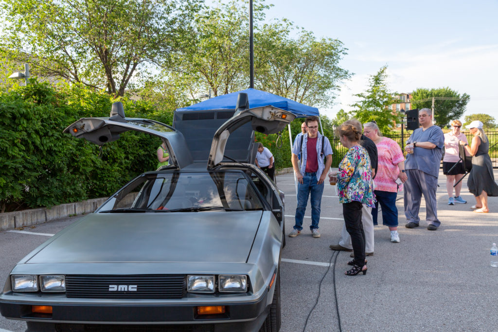 The Delorean sits on the left of the image with its gullwing doors ajar while the owner talks with some drive-in attendees