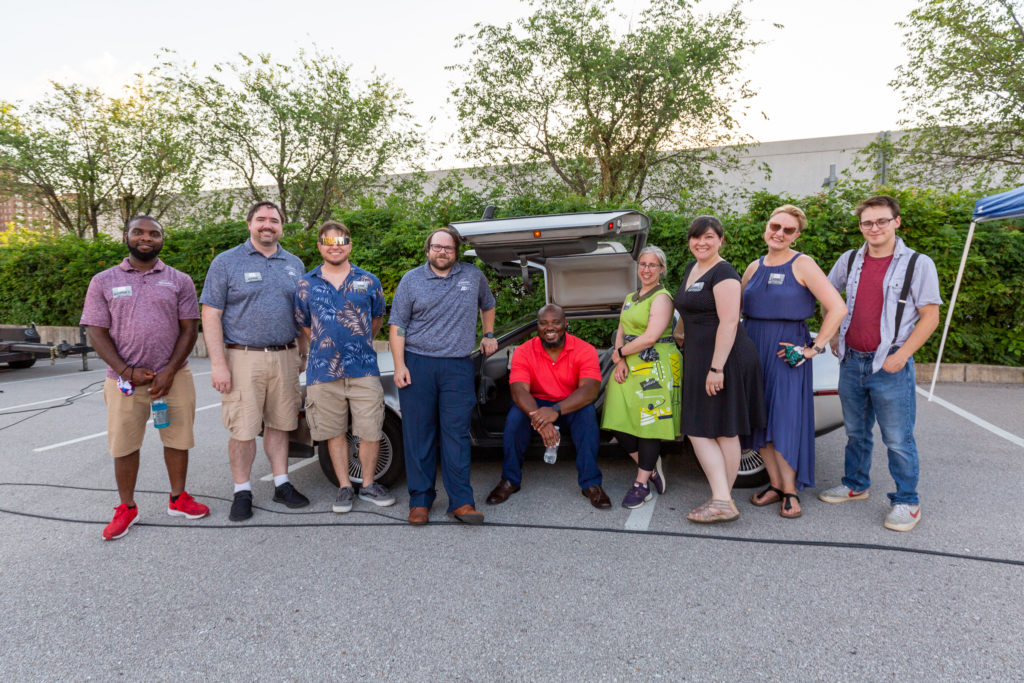 The MindsEye staff pose in front of the Delorean with the Delorean owner. From left to right, Rodney Cotton, Adam Clark, Tom Williams, Mike Curtis, Jason Frazier who is seated in the Delorean, Magan Harms, Angela Banks, Laura Foughty, and the Delorean owner