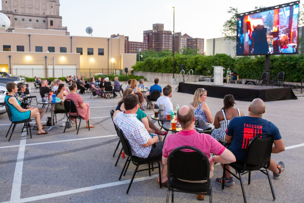 Several groups of people seated around small, round tables enjoy the feature presentation of Back to the Future 2 being played on a large, LED screen