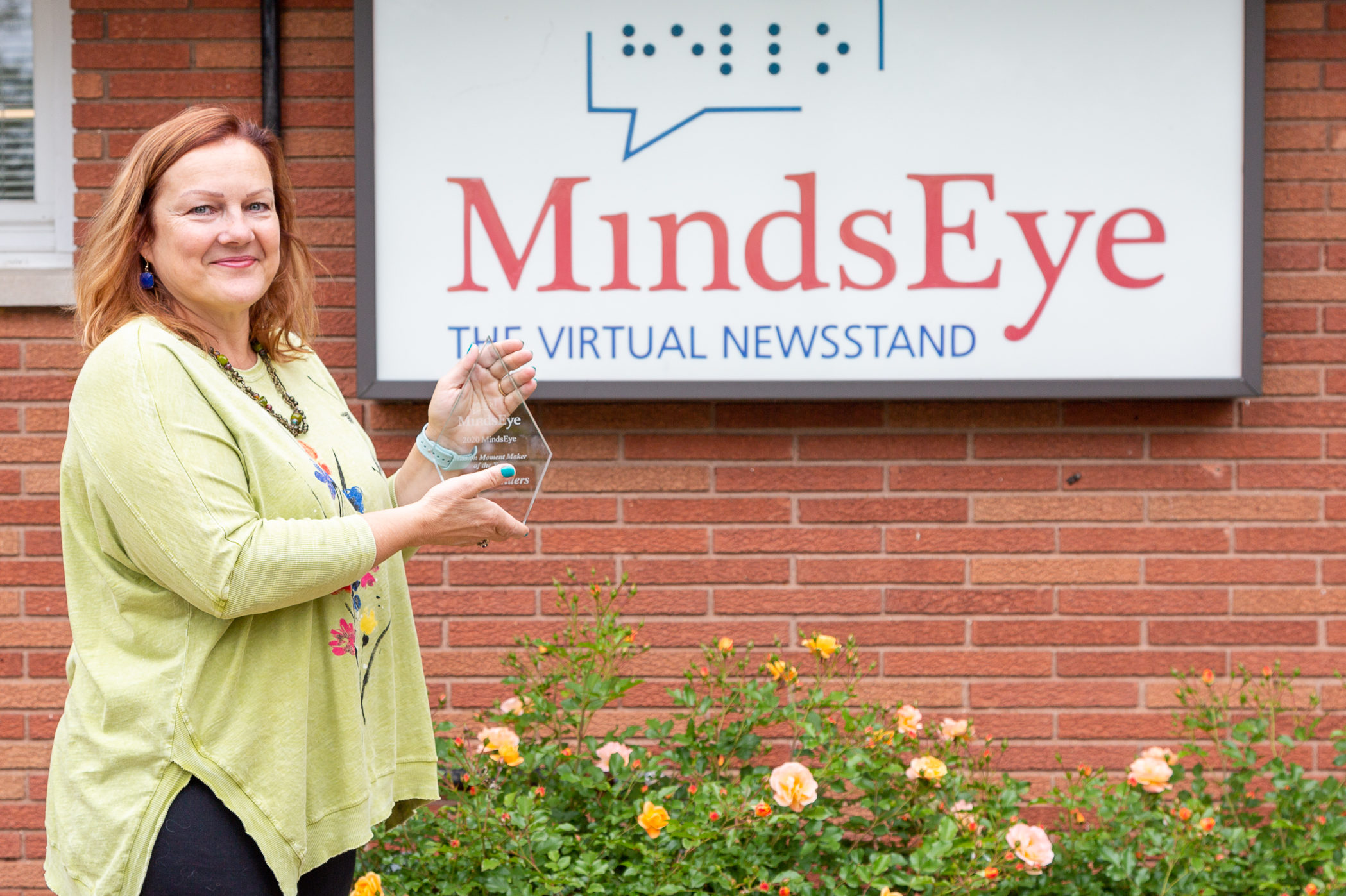 Marge Sanders holds her glass volunteer award and stands in front of the MindsEye studio.
