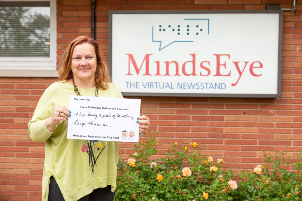 Marge Sanders stands in front of the MindsEye studio holding a sign that says I'm a MindsEye volunteer because I love being a part of something larger than me