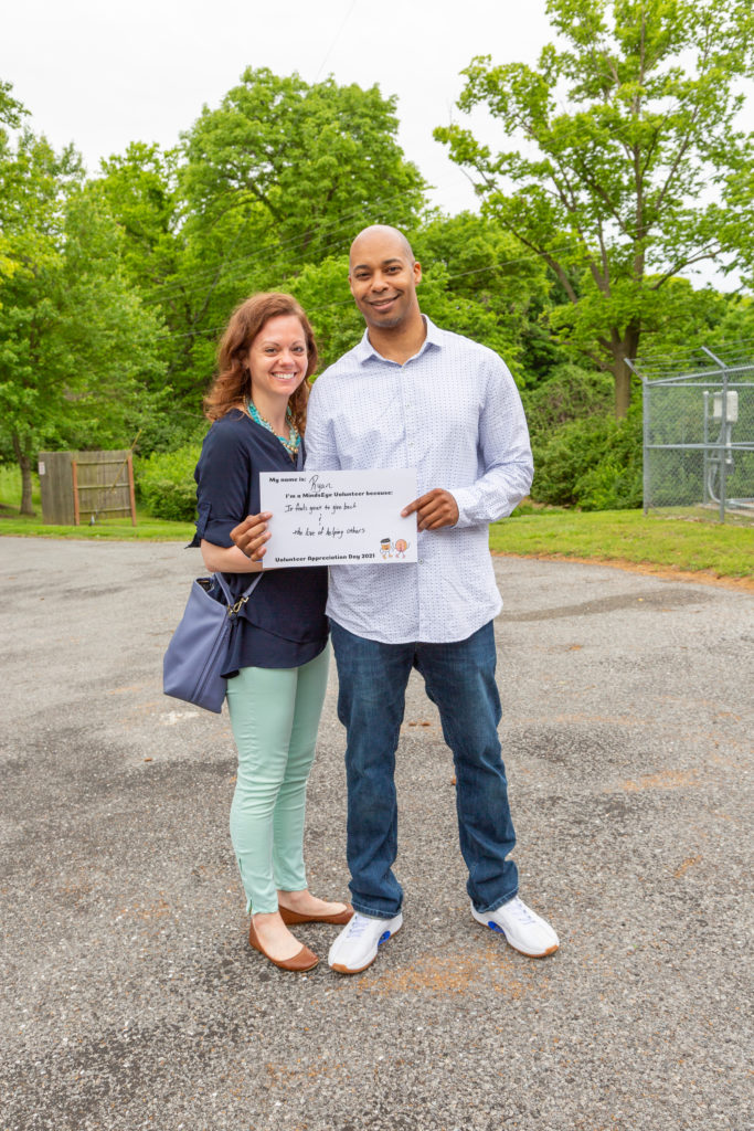 Michelle and Ryan Hune stand in the MindsEye parking lot holding a sign that reads My name is Ryan. I'm a MindsEye volunteer because it feels great to give back and the love of helping others