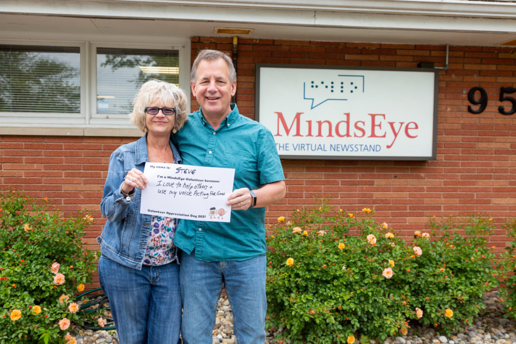 Kathy and Steve Walden stand in front of the MindsEye studio holding a sign that reads My name is Steve. I'm a MindsEye volunteer because I love to help others and use my voice acting for good