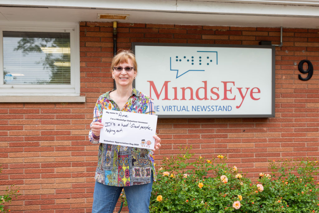 Ame Lemieux stands in front of the MindsEye studio holding a sign that reads My name is Ame. I'm a MindsEye volunteer because It's a hoot! Great people, helping out