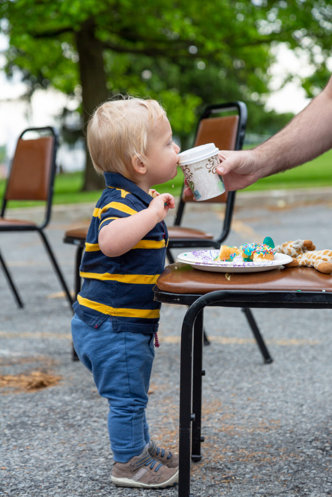 A young boy about two-years-old stands in front of a chair on which there is a paper plate with a partially eaten doughnut on it. The boy is drinking from a cup being held by an adult's hand.