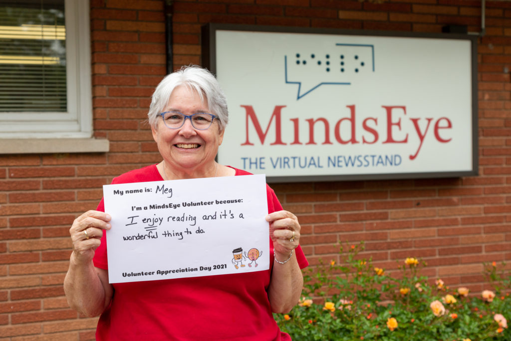 Meg Franklin stands in front of the MindsEye studio holding a sign that reads My name is Meg. I'm a MindsEye volunteer because I enjoy reading and it's a wonderful thing to do.