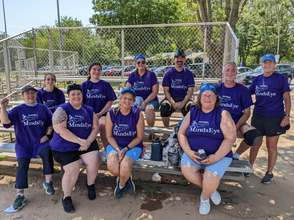 Photo of Lions District 26-M2 BeepBall Team, in matching purple jerseys, sitting in the stands