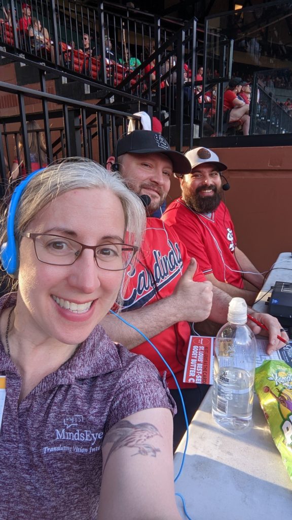 Describers Magan, Dave, and Cory wear headphones and mics at a table with equipment and snacks at Busch Stadium.