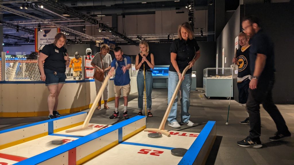 Andrew and Tracey hold shuffleboard-type sticks and push oversized pucks down replica ice.