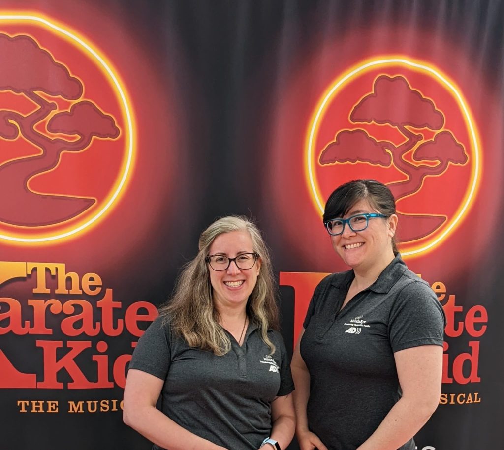 Magan and Angela in front of The Karate Kid The Musical backdrop with a red bonsai inside a golden ring.