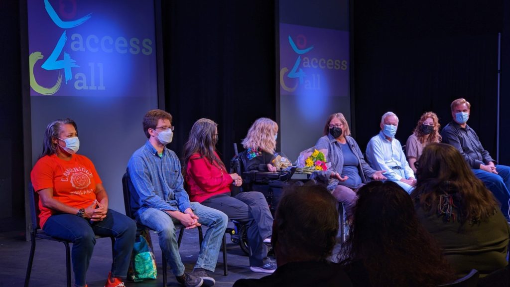 A photo of some of the cast and crew during a post-show talk. Katie sits in the center with a bouquet of flowers on the tray of her power chair. Behind them, large screens say Access4All. 