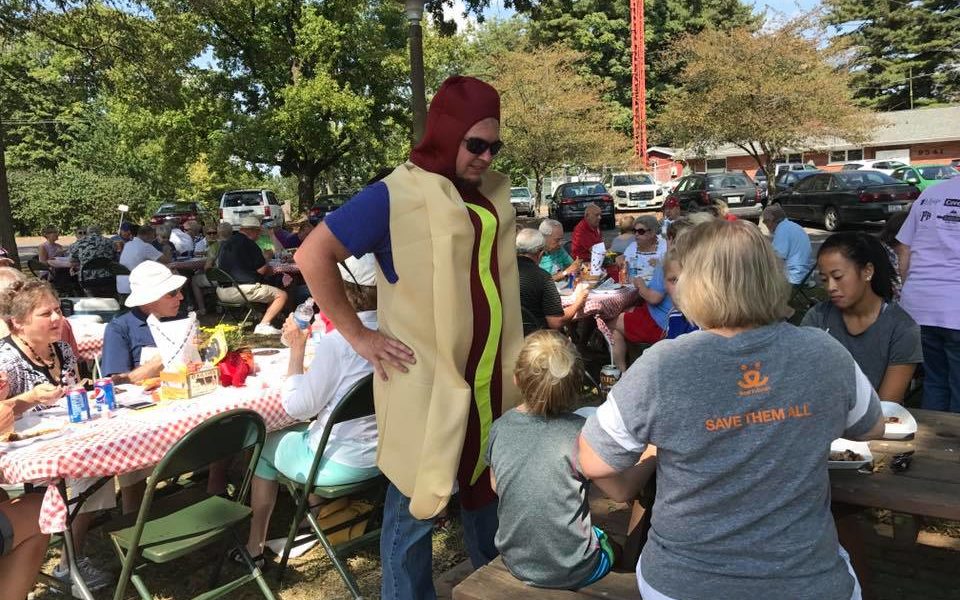 Tom, dressed as a hot dog and wearing dark sunglasses, stands with his hands on his hips as he is talking to a group of people at a wooden picnic table. A group of about 60 volunteers are gathered at 8 rectangle tables covered with red and white checkered tablecloths (adorned with yellow mums and games and bubbles.) They sit in fellowship together, talking and eating smoked chicken and pork steak on a very bright (and warm) September afternoon.