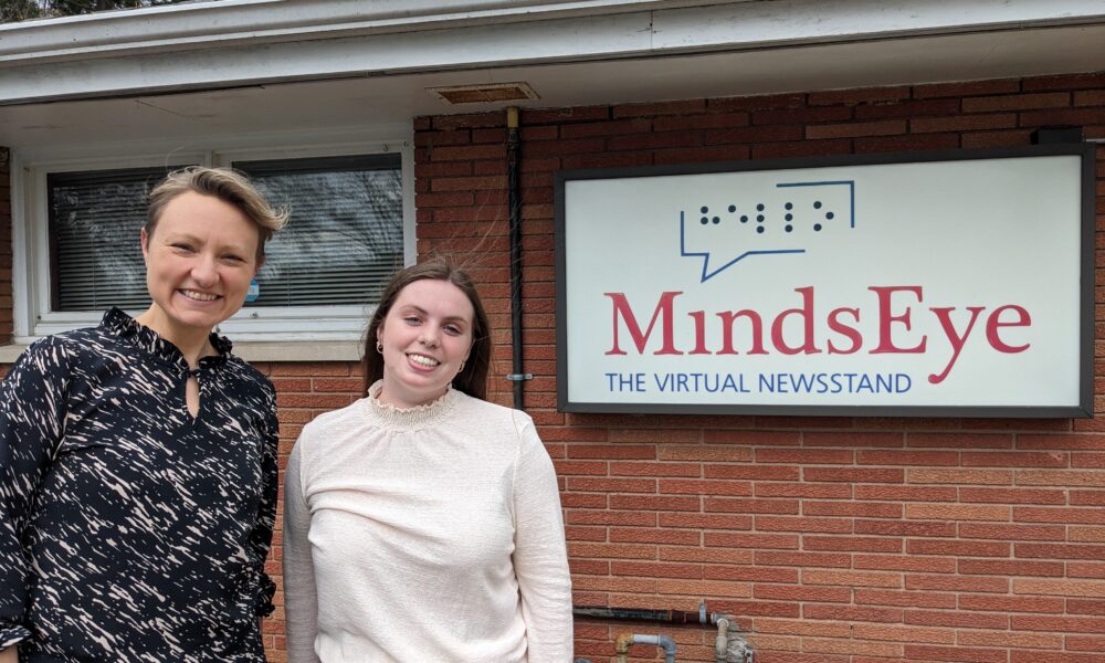 Laura and Skyler smile in front of the MindsEye studio sign, their hair blowing in the wind.