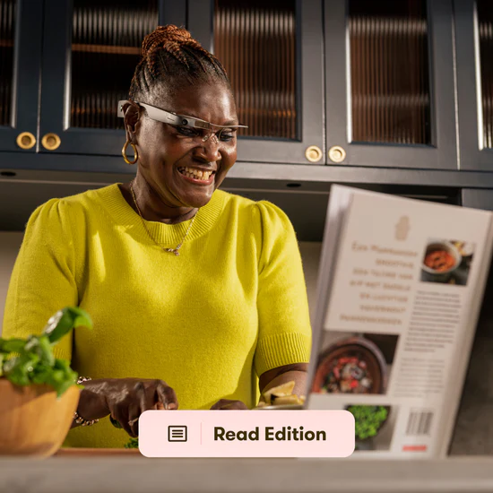 A smiling woman wearing smart glasses reading a cookbook while chopping spices in the kitchen.