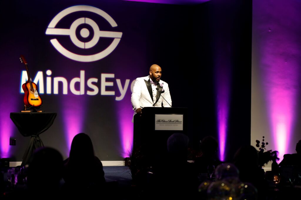 Pascal stands in front of new MindsEye logo, an icon of an eye with a curved line above and below a horizontal line with a circle in the center.